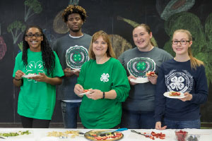 4-H youth and a chaperone smiling around a table with plates of food on it and in their hands.