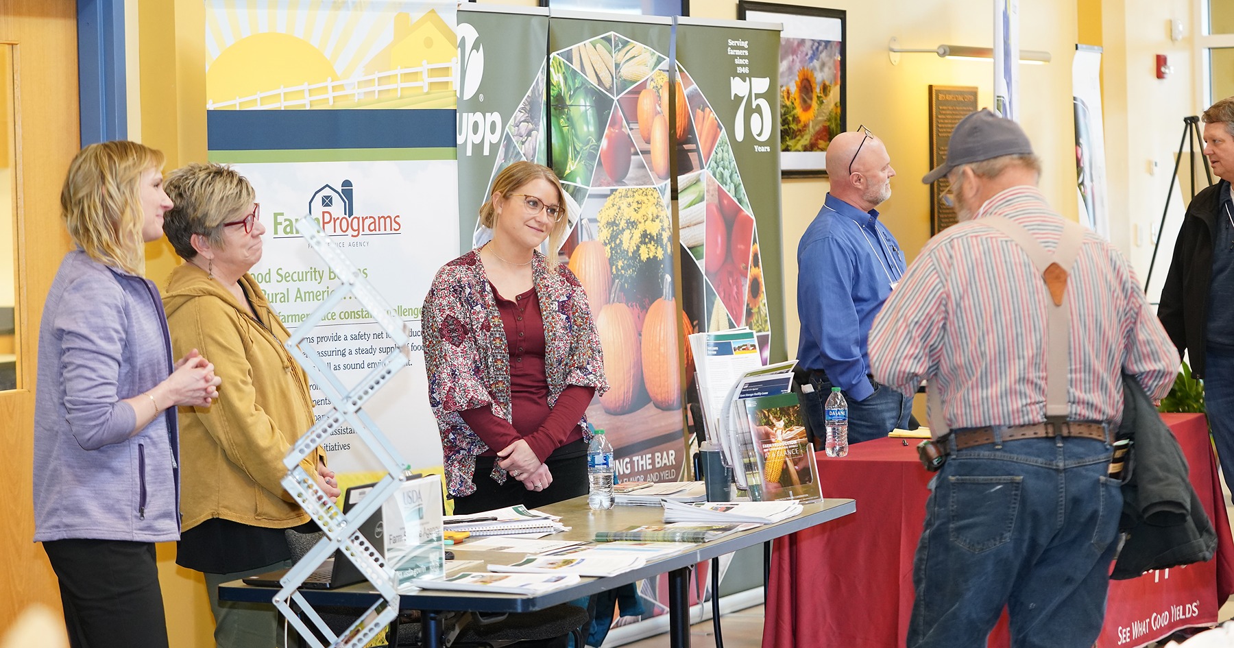 Three women stand behind a table at an agriculture trade show as conference participants pass by.
