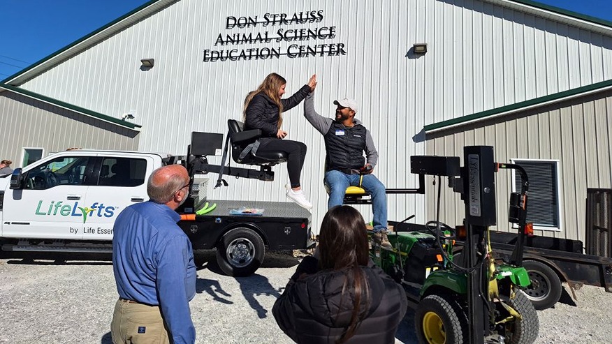A man on a raised tractor seat high-fives a woman seated on a raised seat attached to a pick-up truck.
